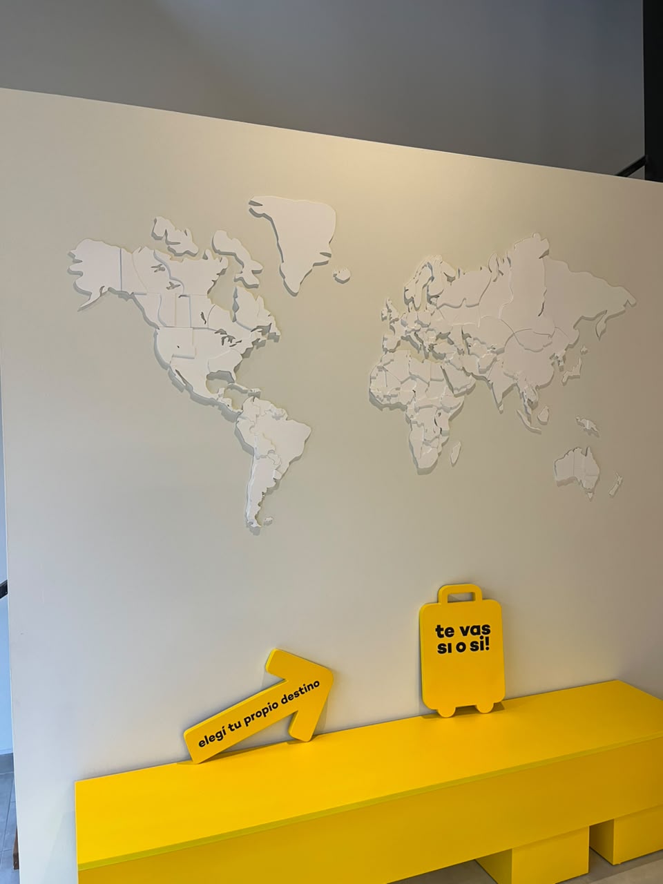 Pared con mapamundi en relieve, banco amarillo y los carteles “Elegí tu propio destino” y “Te vas sí o sí”